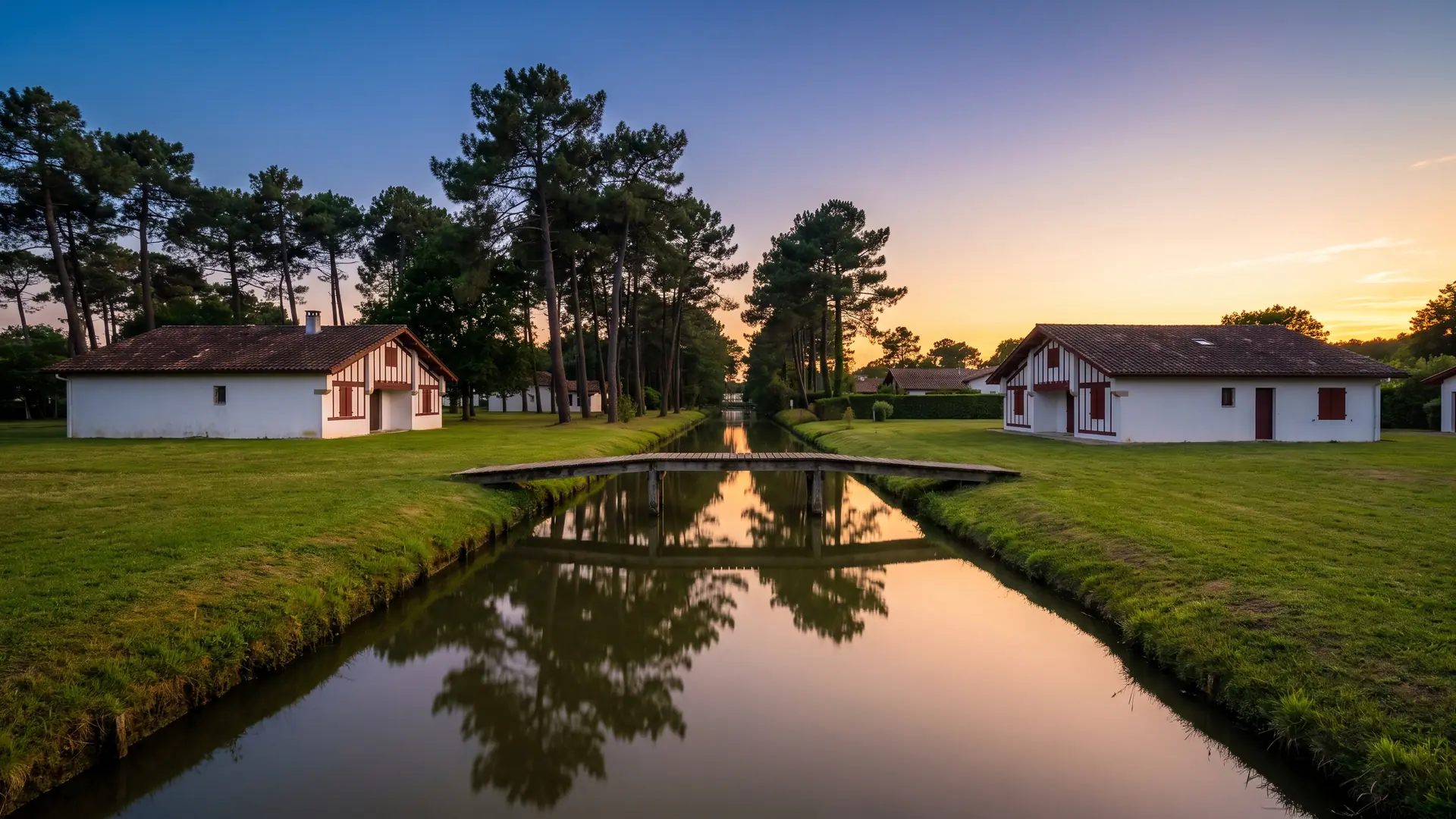 Un canal calme bordé de pins et de maisons landaises à colombages, une passerelle de bois à l'heure dorée