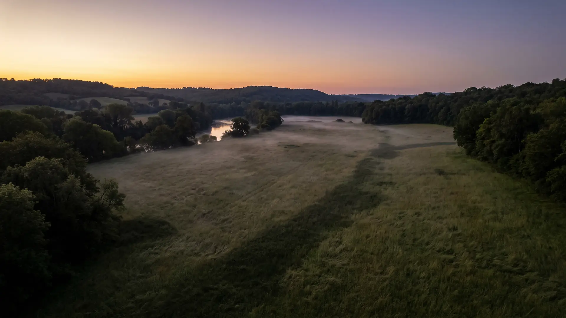 Une vaste prairie au bord de la Dordogne au crépuscule, brumes légères et bois sombres à l'horizon, un paysage solennel