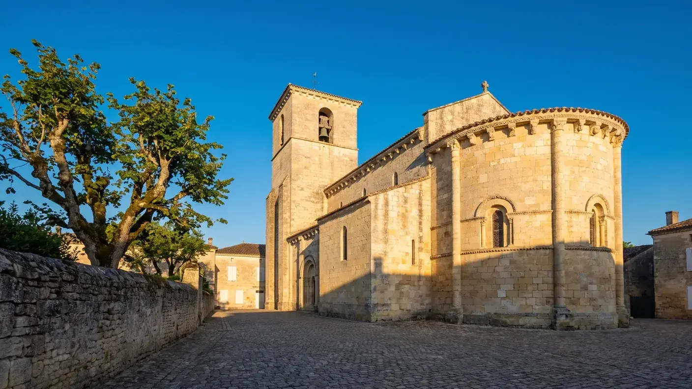 Une église de village en pierre calcaire claire en Gironde, chevet arrondi, clocher carré et place bordée d'un tilleul