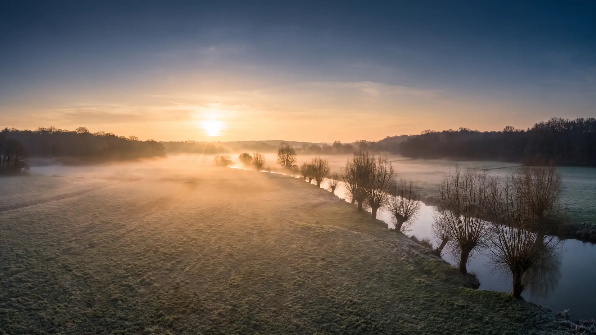 Une vaste prairie humide et une plaine ouverte au bord d'une rivière calme du Libournais en Gironde à l'aube, une brume dorée basse flottant sur l'herbe plate, une ligne d'arbres têtards dénudés et des bois au loin, la première lumière chaude du lever du soleil rasant le champ, une plaine historique solennelle et déserte, ciel doux, absolument aucune personne