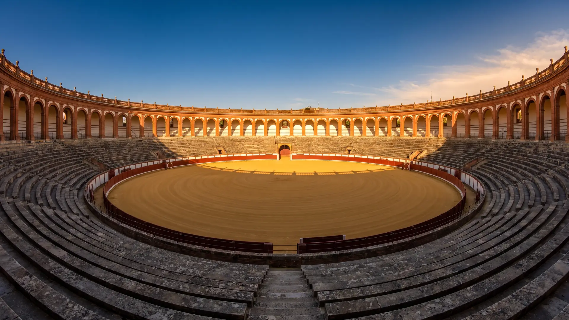 Des arènes historiques vides en brique et arcades blanches, sable doré et gradins déserts à l'heure dorée