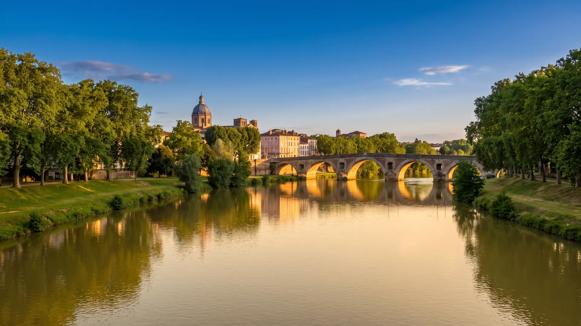 L'Adour calme bordé de berges arborées et d'un pont en arc, façades pastel et dôme au loin, dans les Landes