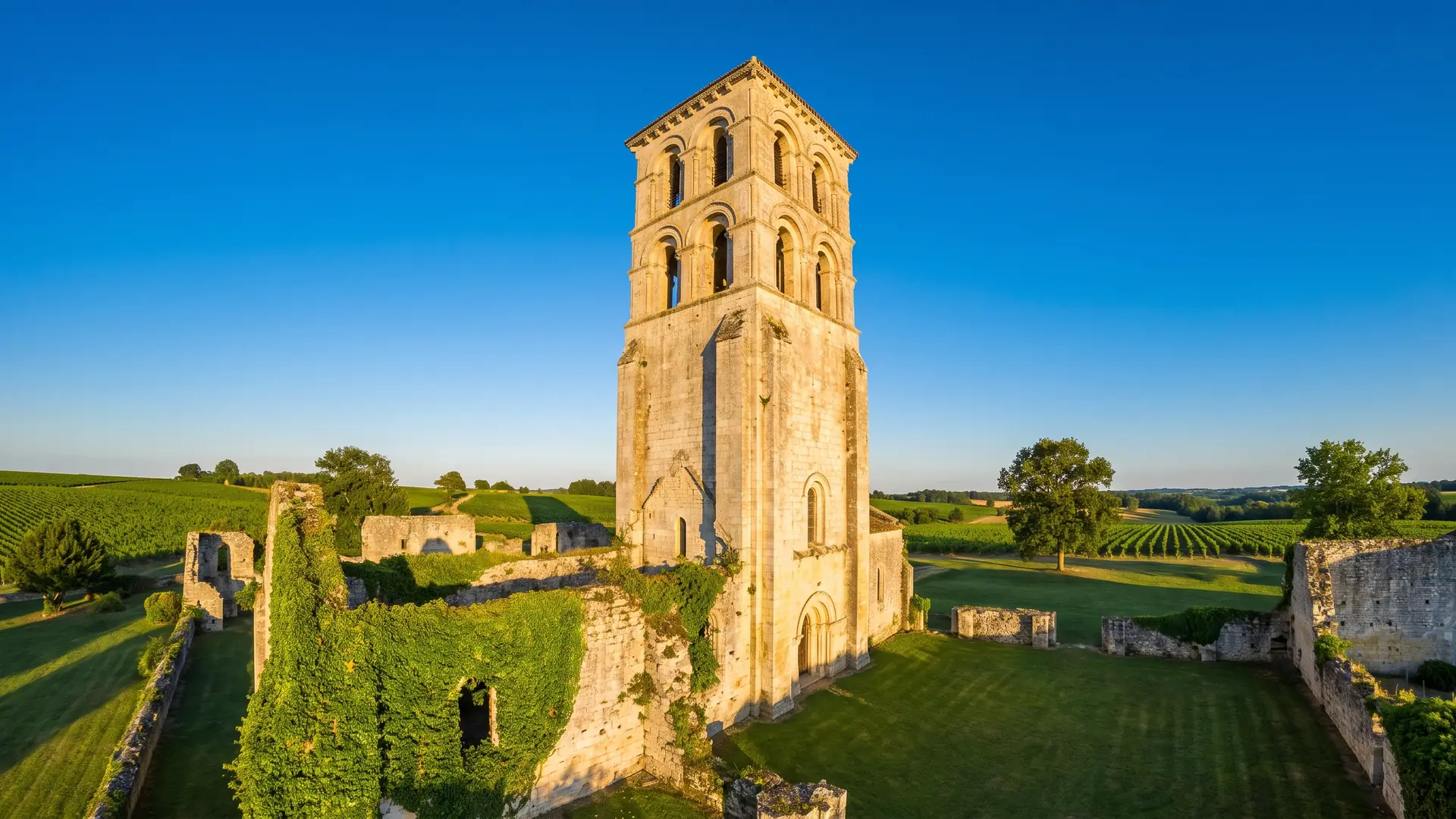 Un haut clocher d'abbaye romane en pierre claire se détachant sur un ciel bleu au-dessus de murs en ruine