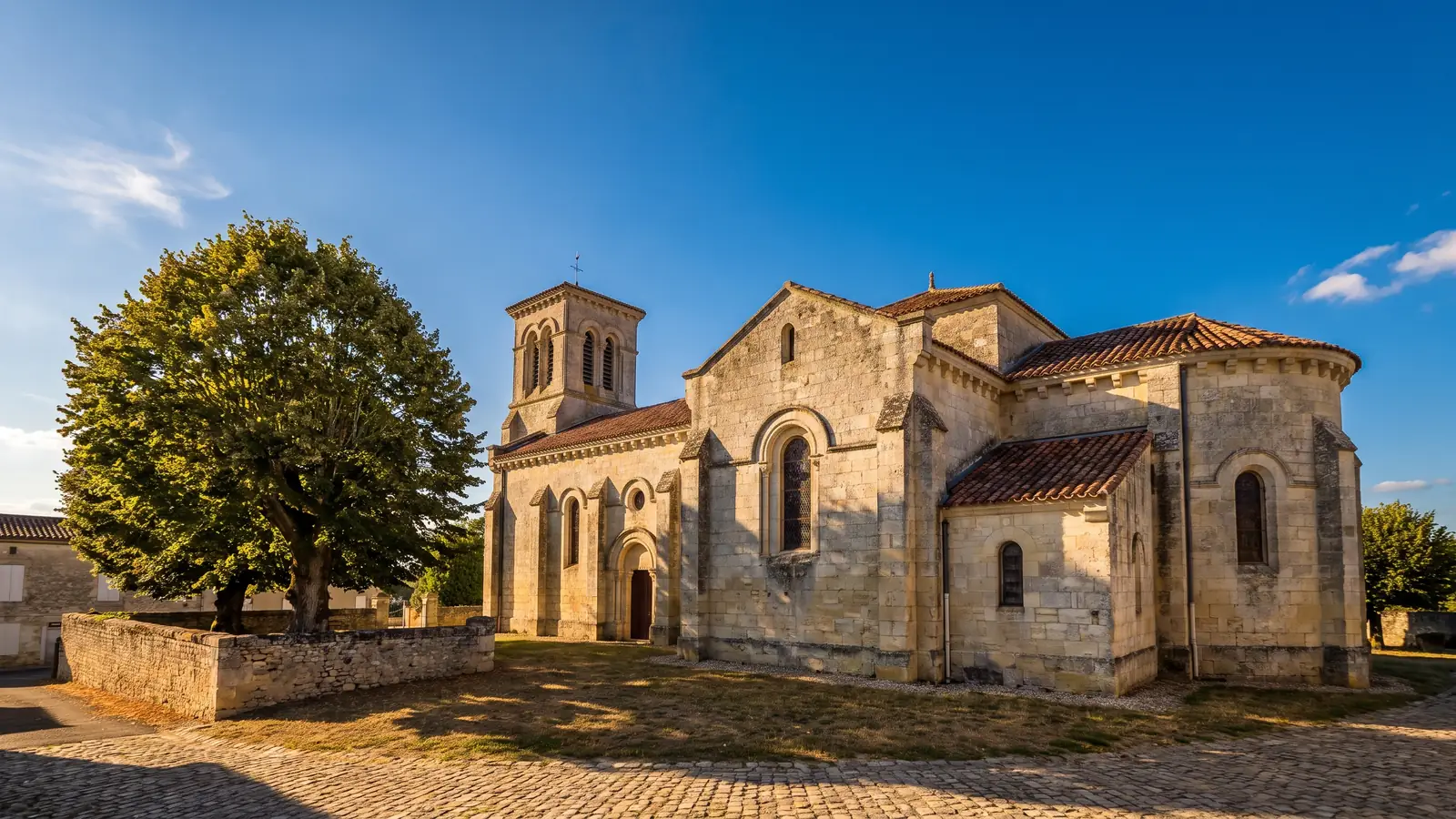 Une église de village en pierre claire du XIXe siècle, à tour-clocher et hautes fenêtres, sur une place bordée d'un tilleul