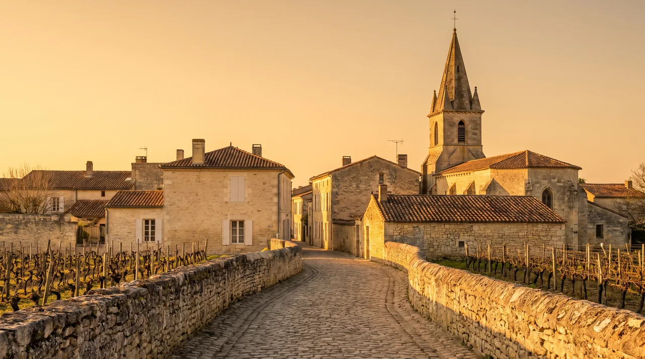 Ruelle tranquille d'un village viticole du Médoc, maisons de pierre claire à volets, clocher au fond, lumière chaude