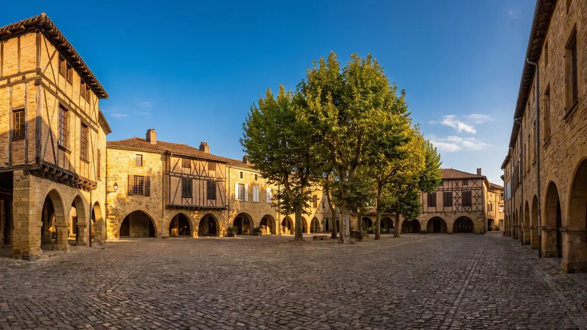 Une place ancienne de Marmande bordée de maisons de pierre et à colombages avec des arcades, toits de tuiles, platanes