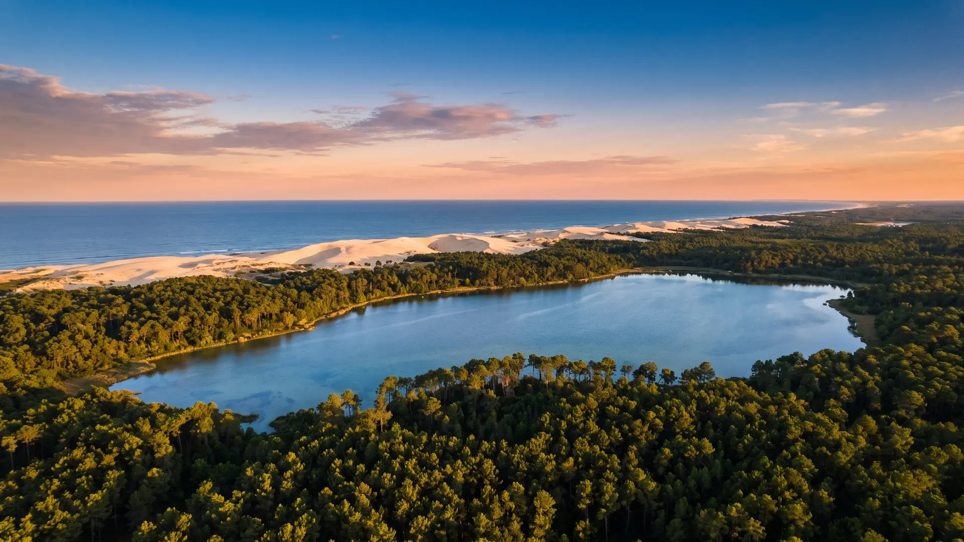 Un grand lac bordé de pins au premier plan, l'immense massif forestier landais puis la ligne des dunes et l'océan au loin