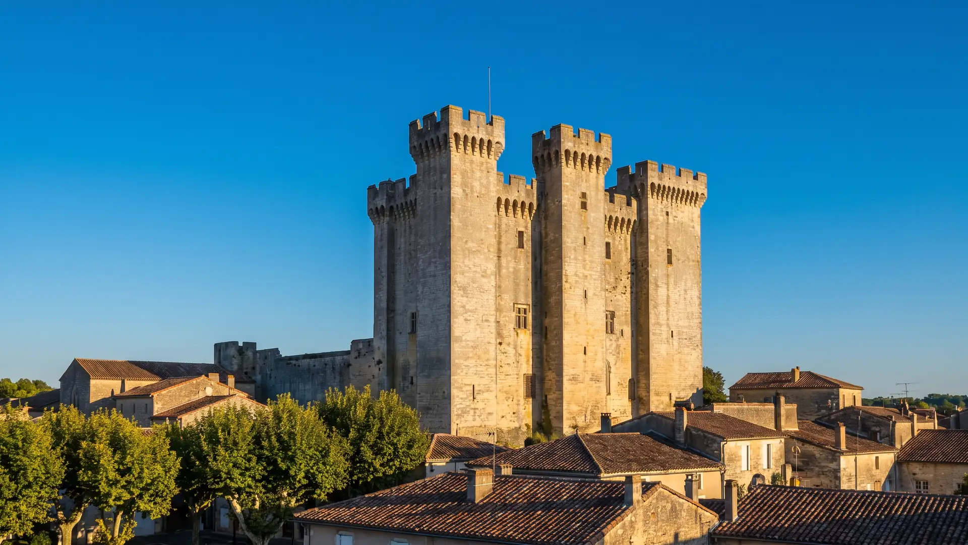 Un donjon médiéval carré à tours jumelles crénelées en pierre gris-doré dominant les toits d'une ville gasconne