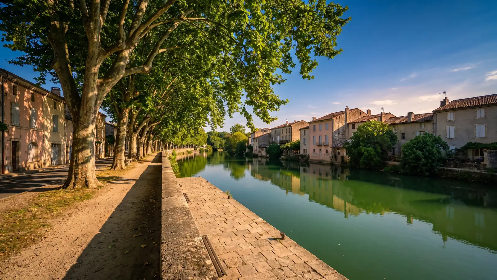 Une promenade ombragée de platanes le long d'une rivière verte, quais de pierre et maisons pastel reflétées