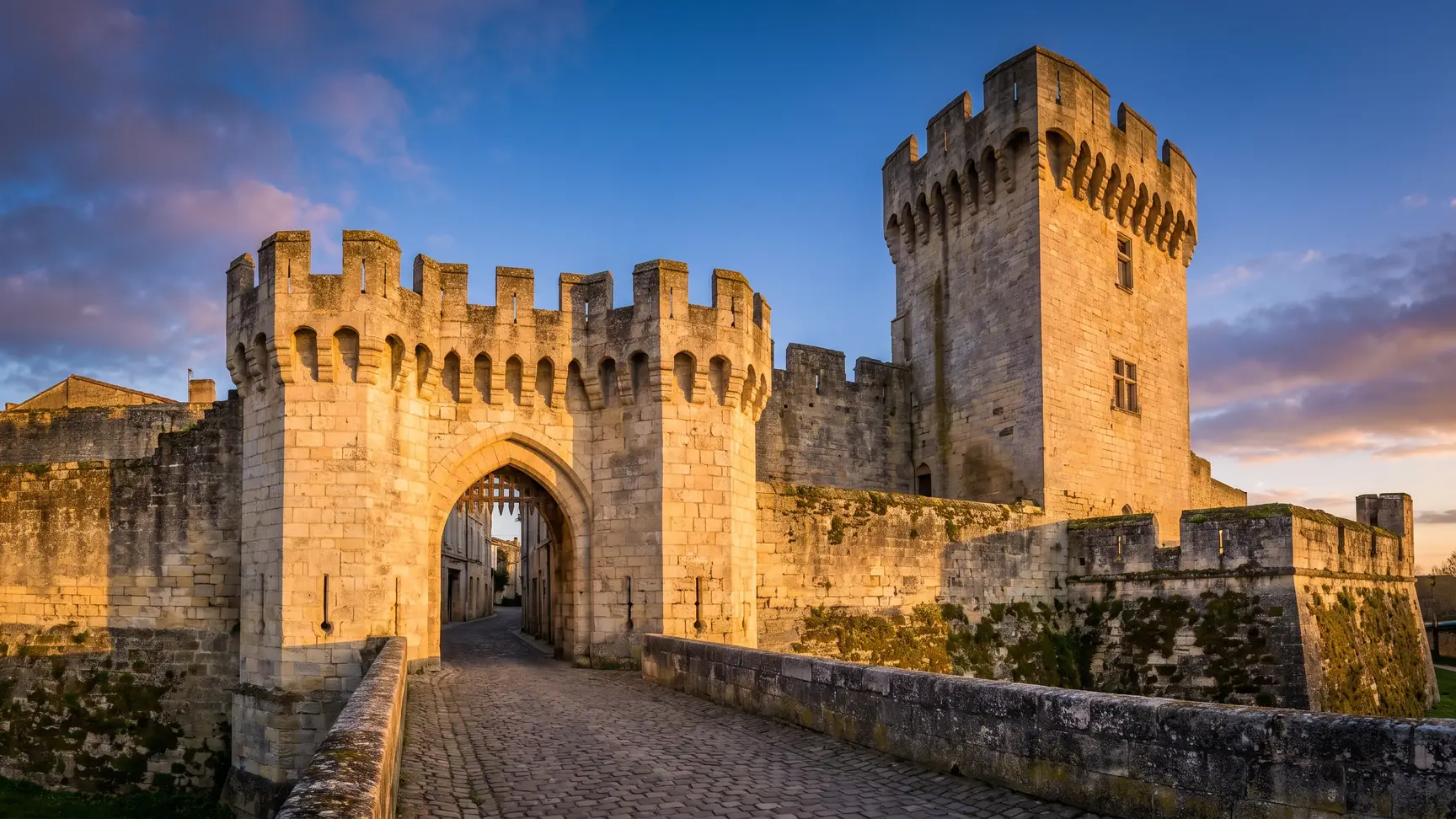Une porte fortifiée médiévale en pierre claire flanquée d'une tour crénelée, à l'entrée d'une cité close