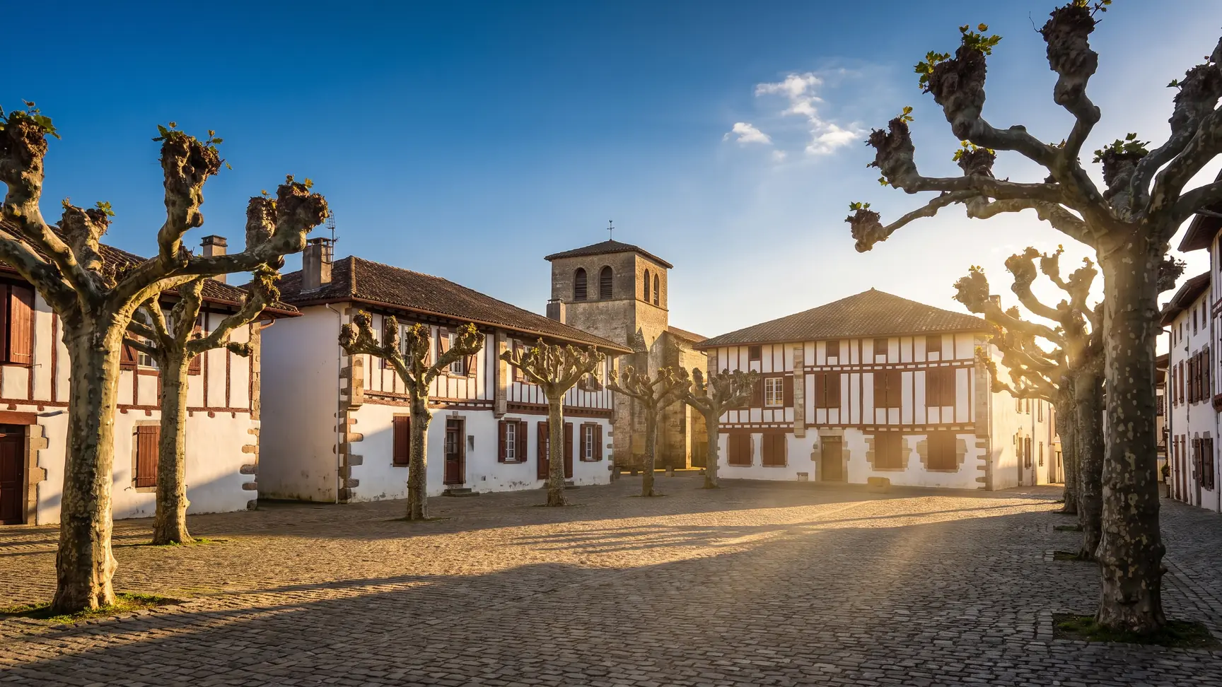 Une place de village landais ombragée de platanes taillés, maisons basses blanches à colombages et clocher d'église en fond