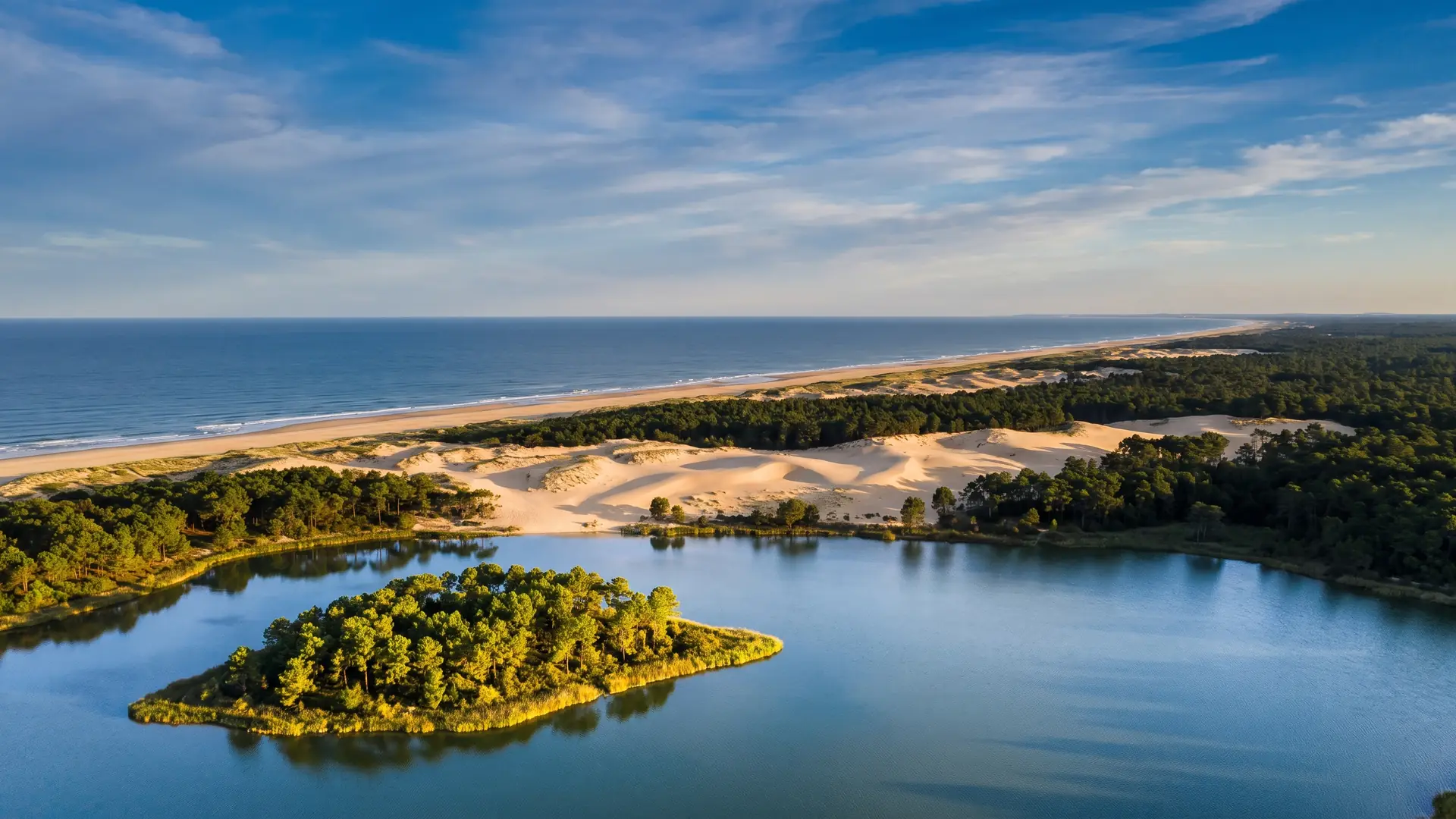 Un panorama du lac marin et de son île boisée au premier plan, dunes, pinède et océan au-delà, sur la côte sud des Landes